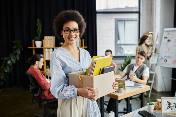 Jolly african american woman packing her items during lay off, colleagues on backdrop.
