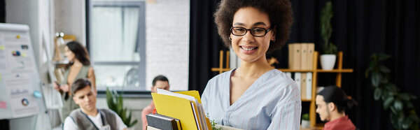 Merry african american woman packing her items during lay off, colleagues on backdrop.
