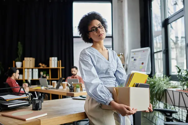 stock image Dedicated african american woman packing her items during lay off, colleagues on backdrop.