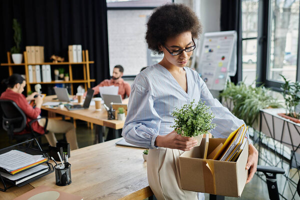 Good looking african american woman packing her items during lay off, colleagues on backdrop.