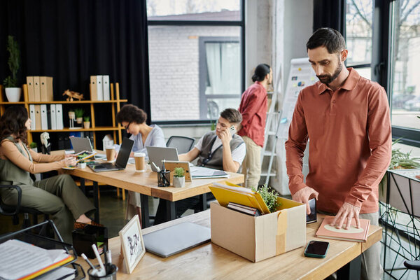Мужчина в повседневной элегантной одежде упаковывает свои вещи во время увольнения, коллеги на заднем плане.