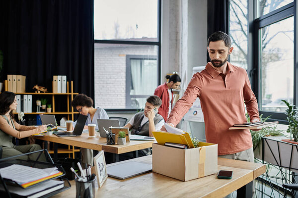 Мужчина в уютной одежде упаковывает свои вещи во время увольнения, коллеги на заднем плане.