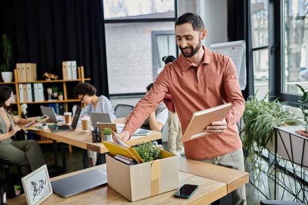Человек с бородой упаковывает свои вещи во время увольнения, коллеги на заднем плане.