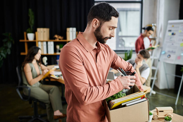 Преданный человек собирает свои вещи во время увольнения в современном офисе, коллеги на заднем плане.