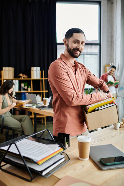 Бородач пакует свои вещи во время увольнения в современном офисе, коллеги на заднем плане.
