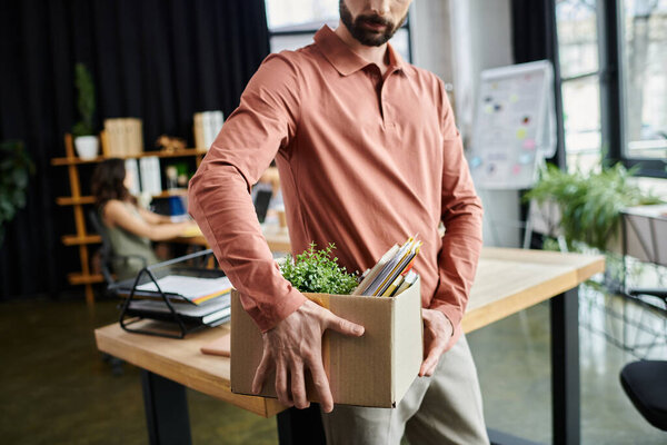 Good looking man packing his belongings during lay off in modern office, colleagues on backdrop.