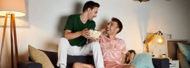 Two men share laughter and popcorn on a cozy night in their living room. clipart