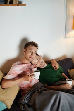 Two men relax together, sipping warm drinks and embracing on a cozy couch. clipart