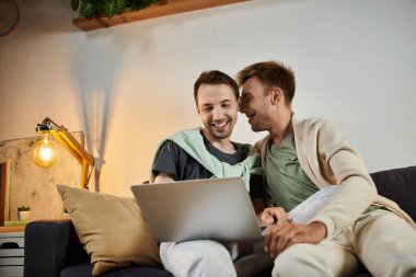 Two men share joyful moments together while using a laptop on their couch. clipart