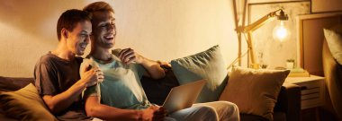 İki adam rahat evlerinde dizüstü bilgisayarda çalışırken kahkahalarını paylaşıyor..