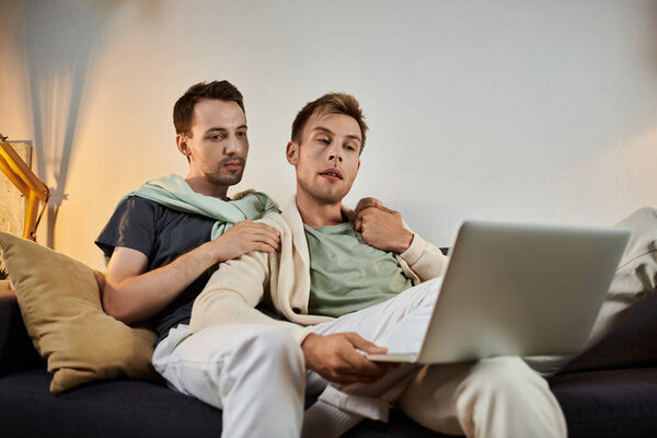 Двое мужчин разделяют нежный момент во время отдыха на диване с ноутбуком.