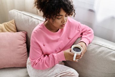 A young woman sips coffee while relaxing on the couch in her stylish living room. clipart