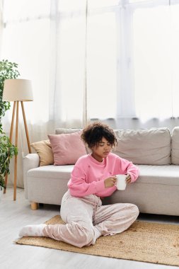 A young woman relaxes on the floor with a cup, soaking in the comfort of her home. clipart