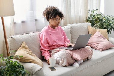 Bir kadın güneşli bir oturma odasında laptopuna odaklanırken kahvesini yudumluyor..