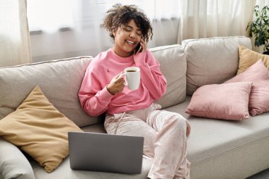 A young woman laughs on the couch while sipping her favorite beverage. clipart