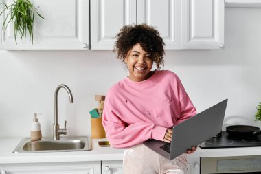 Cheerful woman engages with a laptop while enjoying her cozy kitchen. clipart