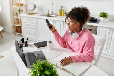 Genç bir kadın kahvesini yudumlarken ve telefonunu kontrol ederken ders çalışmaktan zevk alıyor..
