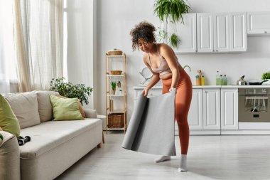 Bir kadın pırıl pırıl yaşam alanında dinlenirken yoga minderi serer..