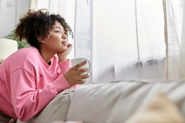 stock image A young woman relaxes on her couch, sipping coffee and lost in thought.