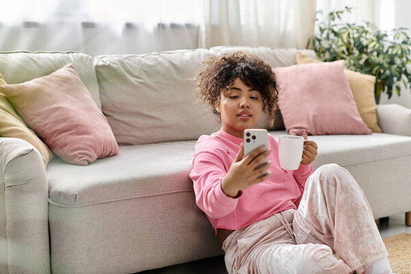 Молодая женщина расслабляется дома, потягивая чай и просматривая свой телефон.