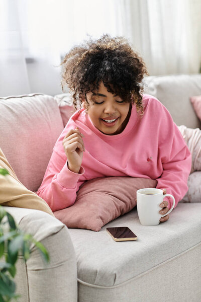 A beautiful woman relaxes on her sofa, sipping coffee and using her phone.