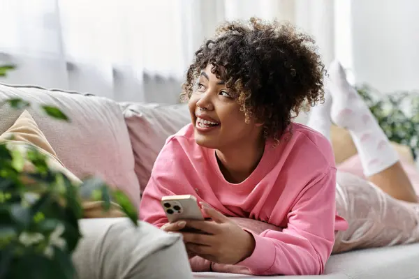 stock image A cheerful young woman relaxes on the couch with her smartphone.