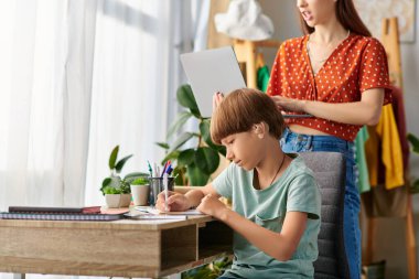 A mother watches her hearing impaired son as he draws creatively at a cozy desk. clipart