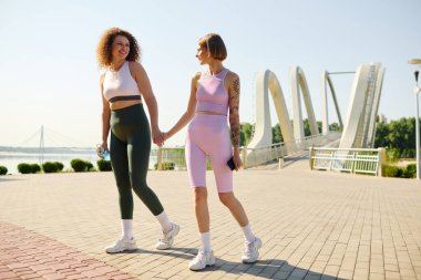 Parkta aydınlık bir günde, iki kadın el ele yürür, gülümserler..