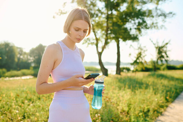 A young woman in a park checks her phone while holding a water bottle.