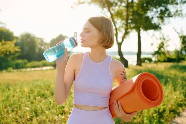 stock image A woman sips water, ready for a yoga session in a vibrant park.