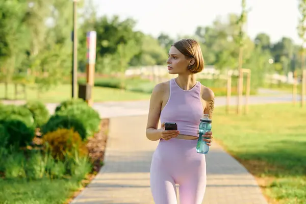 stock image A young woman strolls through a lush park, phone and water in hand.