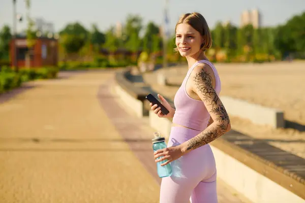 stock image A cheerful young woman walks through a vibrant park with a water bottle in hand.