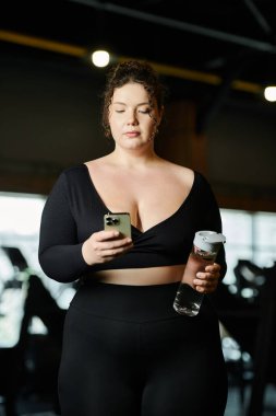 Büyük beden bir kadın spor salonunda biraz soluklanır, suyunu yudumlar ve telefonunu kullanır..