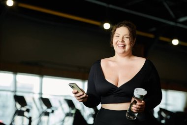 Büyük beden bir kadın spor salonunda su şişesi ve telefonuyla çalışırken kendinden emin bir şekilde gülümsüyor..