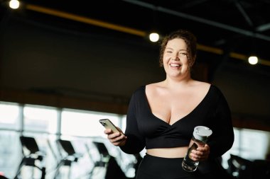 A plus-size woman smiles confidently, engaging in her workout at the gym with a water bottle and phone. clipart