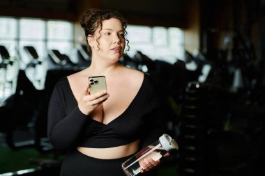 Büyük beden bir kadın telefonunu ve su şişesini tutuyor ve spor salonundaki spor salonuna doğru gidiyor..