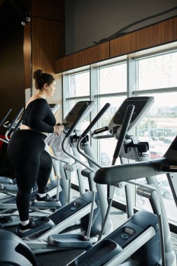 Genç, iri yarı bir kadın, eliptik bir spor salonunda spor yapıyor ve spor yaparken kendine güveniyor..