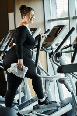 A joyful young woman enjoys her workout on an elliptical, promoting body positivity and wellness. clipart