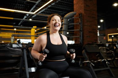 Büyük beden bir kadın coşkuyla spor salonunda halterlerle çalışıyor..