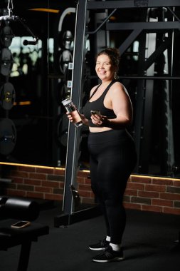 Genç bir kadın spor salonunda vücut pozitifliğini ve gücünü kutlamak için spor kıyafetleriyle neşe içinde egzersiz yapıyor..