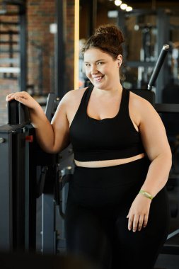 A confident young woman enjoys her workout in activewear, celebrating body positivity in a vibrant gym. clipart