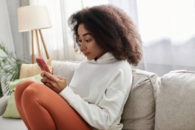 A young woman relaxes on the couch, immersed in her phone while wearing comfy attire. clipart