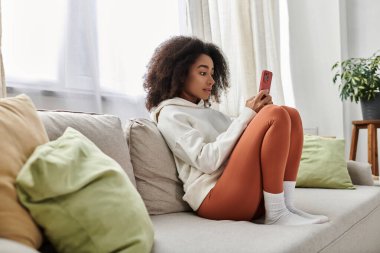 Evde rahat bir an, kapüşonlu ve taytlı genç bir kadın telefonunun keyfini çıkarıyor..