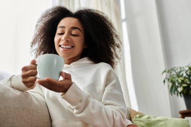 A cheerful young woman in a cozy outfit sips her drink while relaxing at home in her living room. clipart