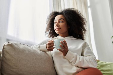 A young woman relaxes at home, savoring a warm drink while wrapped in comfort on her couch. clipart