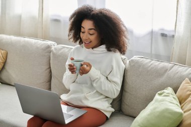 A young woman enjoys her tea, comfortably lounging on the couch while using her laptop at home. clipart