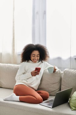 Genç bir kadın evinde dinleniyor, çay yudumluyor ve etrafını rahatlıkla sarmışken telefonunu kullanıyor..