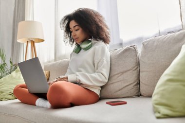 Genç bir kadın evinde rahat bir şekilde koltukta otururken dizüstü bilgisayarıyla kulaklıklarını takıyor..