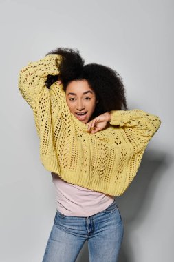 Kıvırcık saçlı genç bir kadın rahat sarı bir kazak giyerken neşeli bir poz verir..