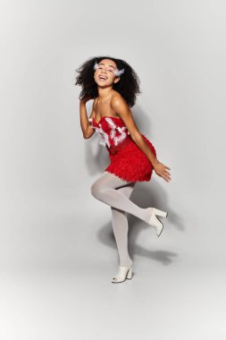 Kıvırcık saçlı genç bir kadın zarif kırmızı bir elbise içinde poz veriyor, neşeli ruhunu sergiliyor..